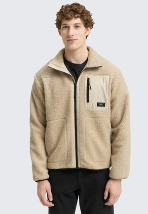 Beige fleece jacket with a high collar, zip front, and a cream pocket featuring black zipper. Soft texture and two front pockets.
