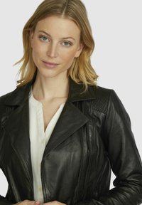 Black leather jacket with ribbed shoulder accents, notched collar, and zip details, worn over a white button-up shirt. Smooth texture.