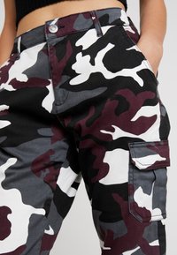 Pantalon cargo camouflage présentant un mélange de motifs noir, blanc, gris et marron. Grande poche latérale avec rabat. Tissu en coton.