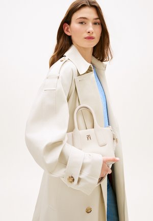 Borsa beige dalla forma strutturata, con manici superiori e dettaglio logo oro, tenuta da una modella con un trench beige abbinato su uno sfondo neutro.