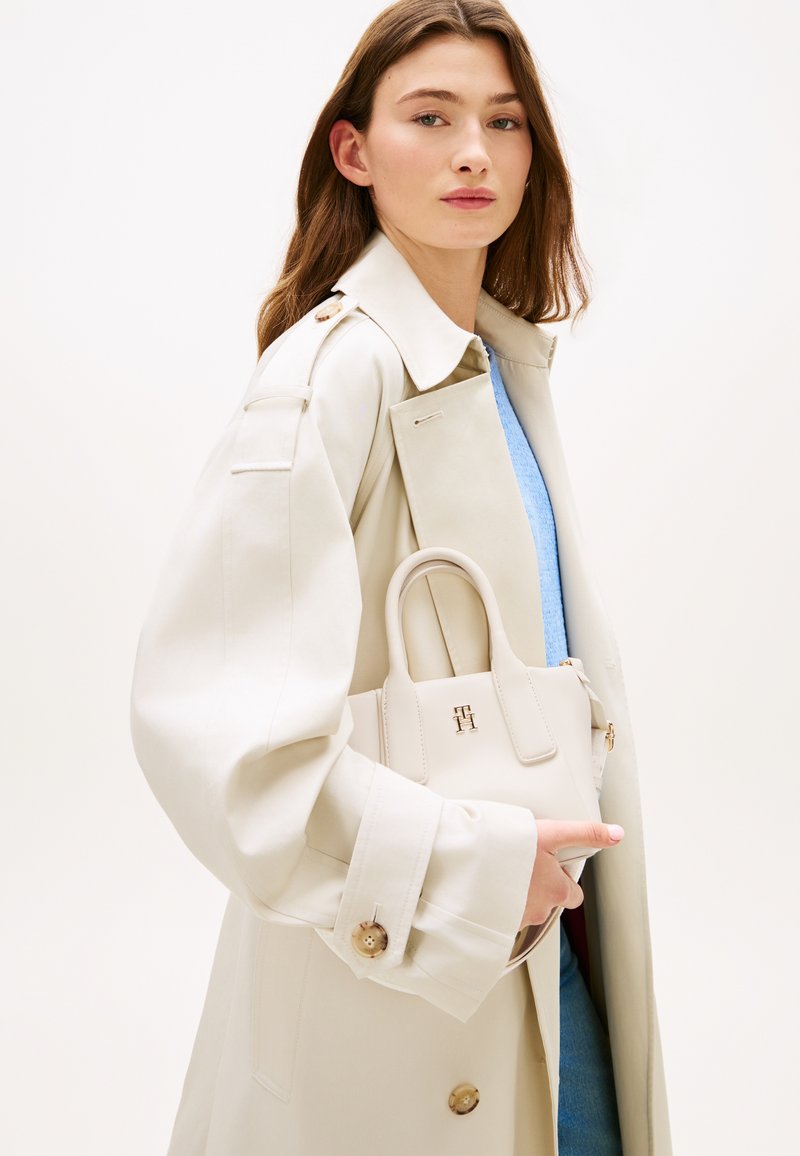 Μπεζ τσάντα με δομημένο σχήμα, χειρολαβές στην κορυφή και χρυσή διακοσμητική λεπτομέρεια με λογότυπο, κρατημένη από μοντέλο που φοράει τζιάνγκλ παιδικό μπεζ σε ουδέτερο φόντο.