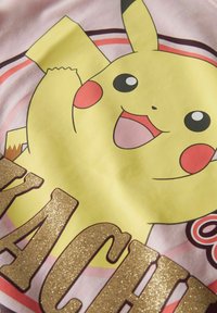 Kollane Pikachu tegel nnäo ja punaste põskedega roosa kangal, mille all on osaliselt nähtav sädelev kuldne tekst.