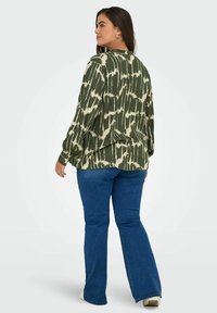 Zelená blůza s béžovým tie-dye vzorem, dlouhými rukávy a volným stříhem, kombinovaná s modrými zvonovými džíny. Jednoduchý kulatý výstřih a splývavá látka.