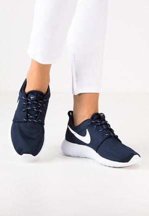 Persoon draagt marineblauwe Nike-sneakers met witte swoosh en zolen, gecombineerd met witte cropped broek tegen een witte achtergrond.
