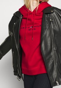 Černá kožená motocyklová bunda přes červenou mikinu s kapucí, která má šňůrky a vyšívané logo značky. Hladké textury, přiléhavý střih.