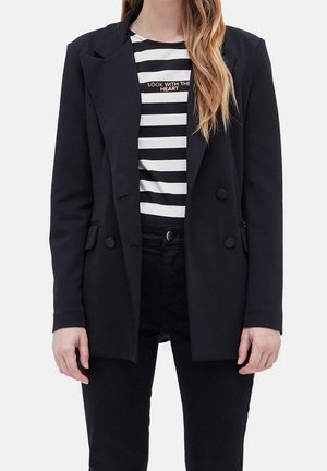 Femme portant un blazer noir croisé sur une chemise rayée noir et blanc avec le texte « LOOK WITH THE HEART », et un pantalon noir.