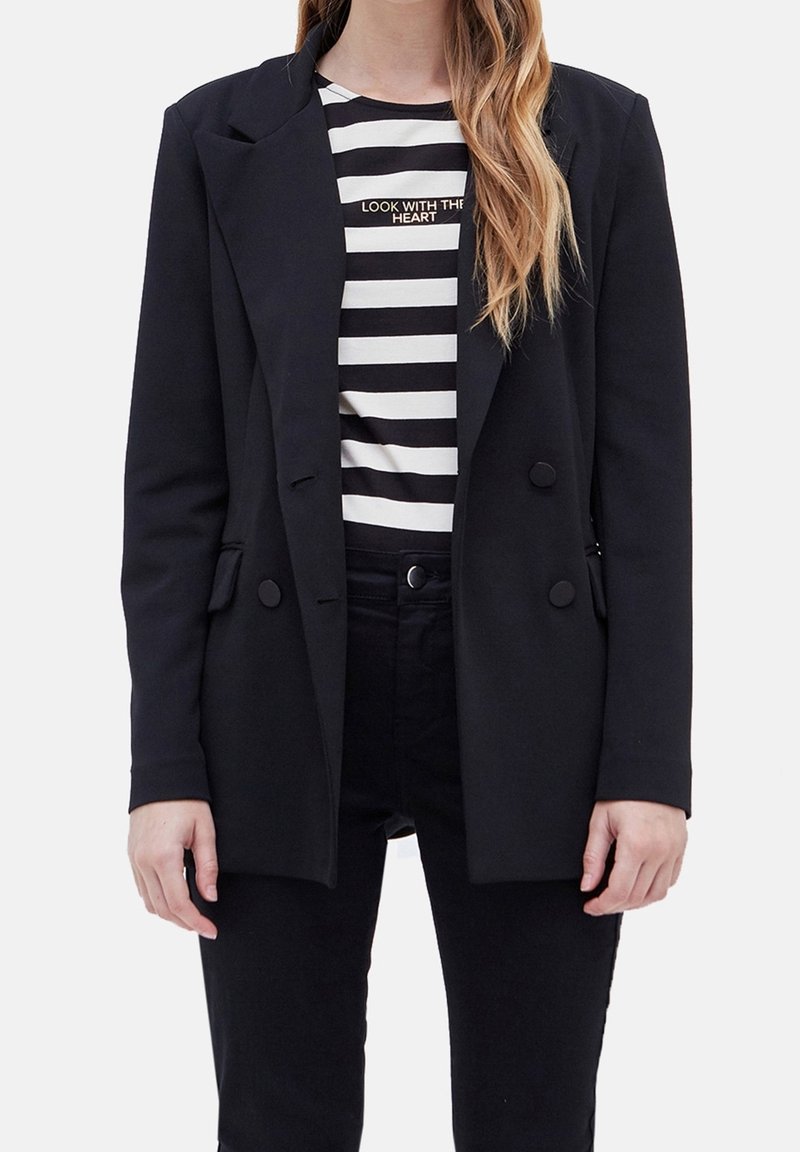 Femme portant un blazer noir croisé sur une chemise rayée noir et blanc avec le texte « LOOK WITH THE HEART », et un pantalon noir.