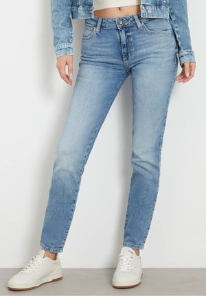 Osoba ubrana w jasnoniebieskie obcisłe dżinsy, krótką kurtkę dżinsową i białe trampki, stojąca na tle białej ściany.