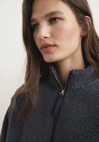 Giacca zip-up in fleece grigio scuro con collo alto e superficie testurizzata. Presenta una zip frontale e un aspetto morbido e accogliente.