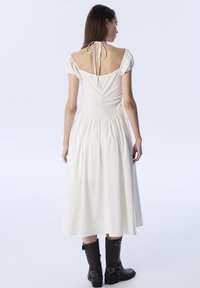 Vestido blanco de longitud media con mangas abullonadas, cintura fruncida y espalda descubierta atada con una cinta; combinado con botines de cuero negro hasta el tobillo.