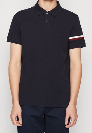 Mann trägt ein marineblaues kurzärmeliges Poloshirt mit einem weißen und roten Streifen am linken Ärmel und einem kleinen Logo auf der Brust.