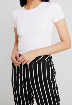 Woman wearing a fitted white ribbed short-sleeve top and black high-waisted pants with vertical white stripes, hands adjusting waistband.