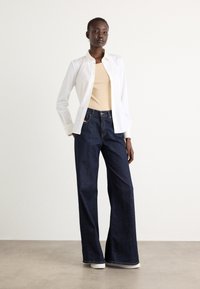 Weißes Hemd über einem beigen Top, kombiniert mit dunkelblauen weiten Jeans. Das Outfit zeichnet sich durch klare Linien und ein minimales Design aus.