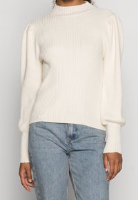 Prążkowany, kości słoniowej sweter z wysokim kołnierzem i bufkami; w połączeniu z jasnoniebieskimi dżinsami. Posiada teksturowany materiał i dopasowany krój.