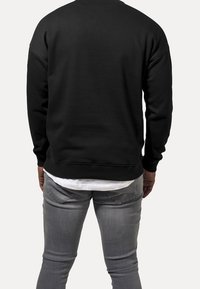 Felpa nera con scollo rotondo e polsini a costine, realizzata in un tessuto morbido. Indossata sopra una camicia bianca e abbinata a jeans grigio chiaro.