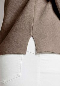 Brauner gestrickter Oberteil mit seitlichen Schlitzen, kombiniert mit weißen Jeans. Die Textur wirkt weich und der Stoff hat ein dezentes Rippmuster.