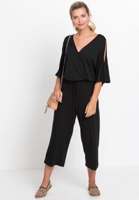 Jumpsuit noir avec décolleté en V, manches courtes avec découpes, taille élastique et pantalons courts, associé à des ballerines à imprimé léopard et un sac à main.