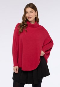 Poncho rosso lavorato a maglia con collo alto, maniche a coste e una forma ampia e fluente, abbinato a una gonna plissettata nera.