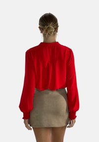 Blusa rossa a maniche lunghe con colletto e polsini arricciati, abbinata a una gonna in suede beige. Texture liscia, vestibilità morbida e dettagli di pieghe sottili sul retro.