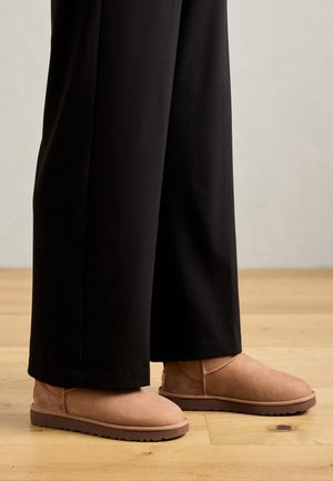 Stivali ankle in suede marrone con suola in gomma piatta, punta rotonda e cuciture minime, indossati con pantaloni neri a gamba larga su un pavimento in legno.