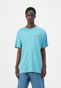 Tirkizna bombažna majica s kratkimi rokavi in krožnim izrezom. Ima oranžni Adidasov logotip na prsih, sproščen kroj in mehak material.