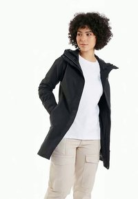 Melna ūdensnecaurlaidīga jaka ar kapuci, ar rāvējslēdzēju un pogu aizdari, sānu kabatām un pieguļošu siluetu. Uzvilkta virs baltas T-kreklu.