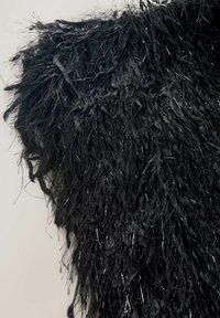 Schwarzes, strukturiertes Kleidungsstück mit langen, zotteligen Fransen, das mit metallischen Akzenten durchzogen ist. Weiches Material mit unterschiedlichen Längen.