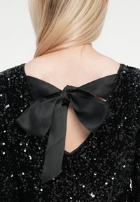 Rochie neagră cu paiete, cu un decolteu adânc în spate, având un funda satinată legată în centru. Textura netedă și strălucirea detaliilor îmbunătățesc designul.