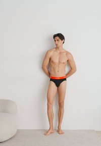 Fekete alsónadrág piros derékkal, fehér színű "Calvin Klein" márkanevével. Simább textúrával, alacsony derékrésszel és testhez simuló fazonnal.