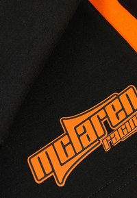 Čierna látka s výrazným oranžovým logom McLaren Racing. Povrch má hladkú štruktúru s kontrastnými oranžovými akcentmi.