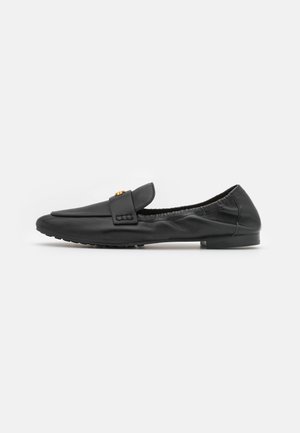 Zapato negro de cuero tipo slip-on con punta redondeada, suela plana y una correa decorativa con un acento dorado. Textura suave y diseño minimalista.