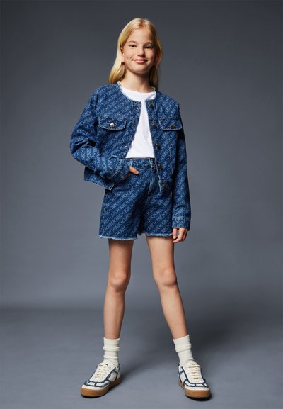 Denim jacket and shorts set in dark blue with all-over logo pattern. White t-shirt underneath and white socks with blue-accented sneakers.