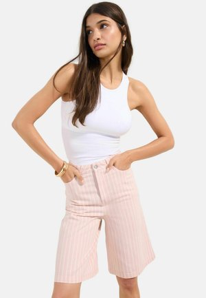 Pruunide pikkade juustega naine, kes kannab valget varrukateta toppi ja kõrge vöökohaga roosade triipudega põlveni ulatuvaid lühikesi pükse, käed taskutes.