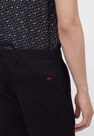 Gros plan d'une personne portant un pantalon noir avec une poche arrière et une chemise noire à motifs rentrée, montrant son bras droit et sa main détendus.
