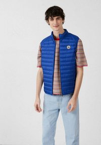 Joven vestido con un chaleco acolchado azul sobre una camiseta de rayas y pantalones vaqueros azul claro, posando frente a un fondo blanco liso.