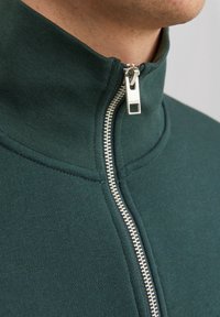 Detailní záběr na osobu v tmavě zeleném zipovém oblečení se stříbrným zipem a vysokým límcem, částečně rozepnutým.