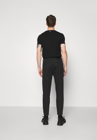 Mężczyzna stojący przodem do białej ściany, ubrany w czarną, dopasowaną koszulkę, ciemne wąskie spodnie, czarne buty, z tatuażem na lewym ramieniu.