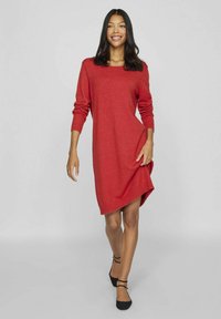 Robe tricotée rouge avec un col rond, des manches longues et une coupe décontractée. Présente un ourlet légèrement asymétrique et des poignets côtelés.
