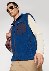 Blauwe fleecvest met een gestructureerd oppervlak, voorzien van een marineblauwe borstzak, ritssluiting en zijzakken. Model draagt een lichtgekleurde lange mouw shirt en een zonnebril.
