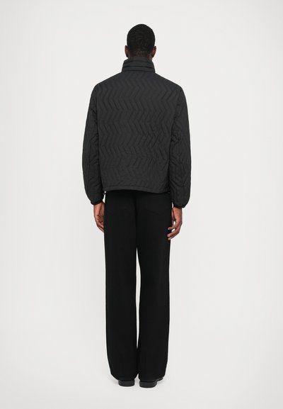 Man wearing a black quilted jacket and black wide-leg pants, standing facing away against a plain white background.