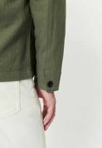 Ruka muža spočívajúca pozdĺž tela, oblečená v olivovo zelenej košeli s dlhým rukávom a čiernym gombíkom na manžete, a v béžových nohaviciach s viditeľnou zadnou vreckom.