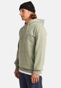 Felpa verde chiaro realizzata in materiale morbido, con tasca frontale, cappuccio con cordino e dettagli del logo sul petto. Indossata con jeans scuri.