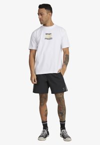 White cotton t-shirt with black and yellow graphic text. Paired with black shorts featuring striped accents and side seams. Black sneakers.