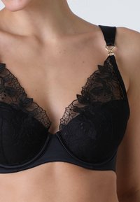 Reggiseno in pizzo nero con motivi floreali lungo il décolleté, corpo in tessuto liscio e spalline regolabili con dettagli in metallo dorato.