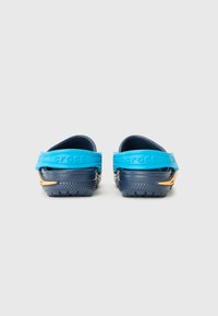 Blå och marinblå crocs med strukturerade ytor, bakremmar och en gul accent. Designen har en klassisk träskotform och gummimaterial.
