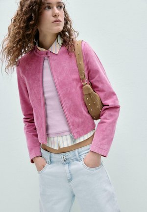 Femme aux cheveux bouclés portant une veste en daim rose sur une chemise rayée et un jean bleu clair, avec un sac à bandoulière marron clouté.