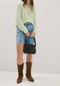 Pull tricot vert menthe, shorts en denim taille haute, bottes hautes en daim marron, et petit sac à main noir avec des accents cloutés.
