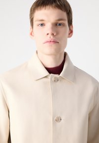 Beige leather jacket with a collar, single button closure, and textured surface; worn over a maroon sweater. Simple, modern design.