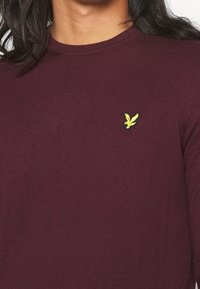 Bordeauxroter Langarmpullover aus weichem Stoff, mit einem gerippten Kragen und einem kleinen gelben, gestickten Vogel-Logo auf der Brust.