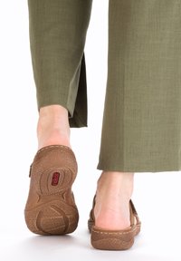 Brown slip-on clogs with a textured outsole, round toe, and decorative stitching. Worn with olive green wide-leg pants.